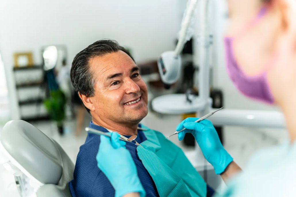 Dentist in teal gloves examines smiling senior male patient in dental chair.