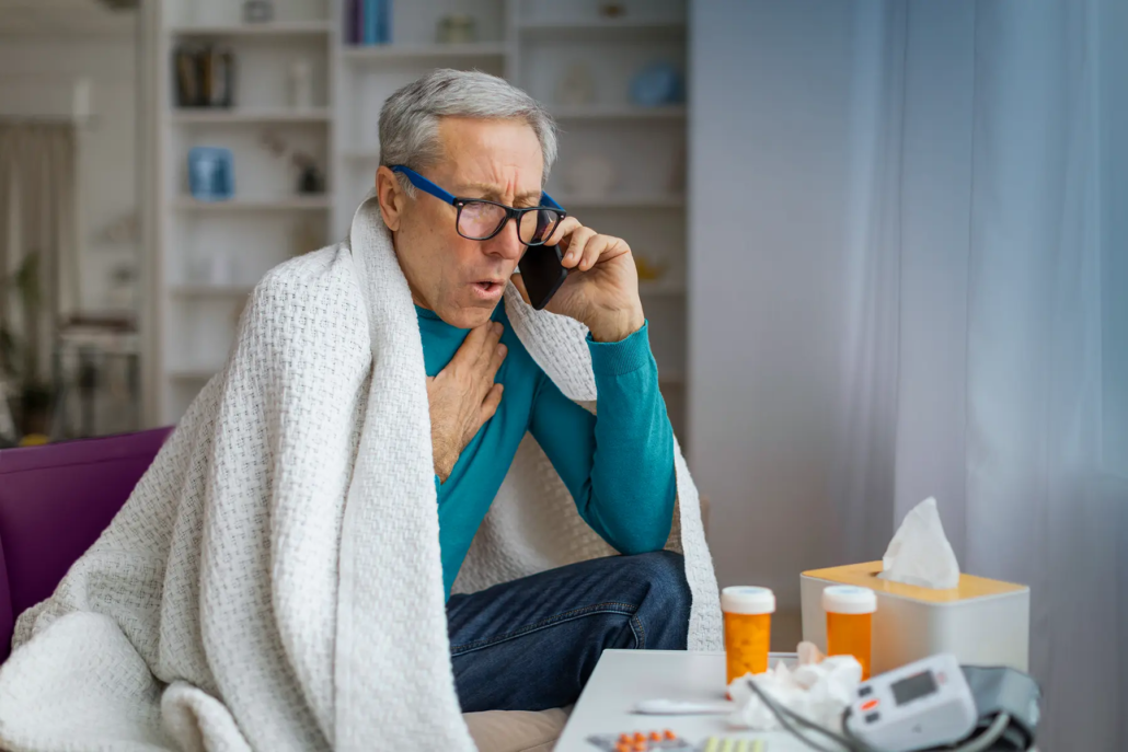 Senior man wrapped in a blanket, wearing glasses, talks on the phone, holding his chest.