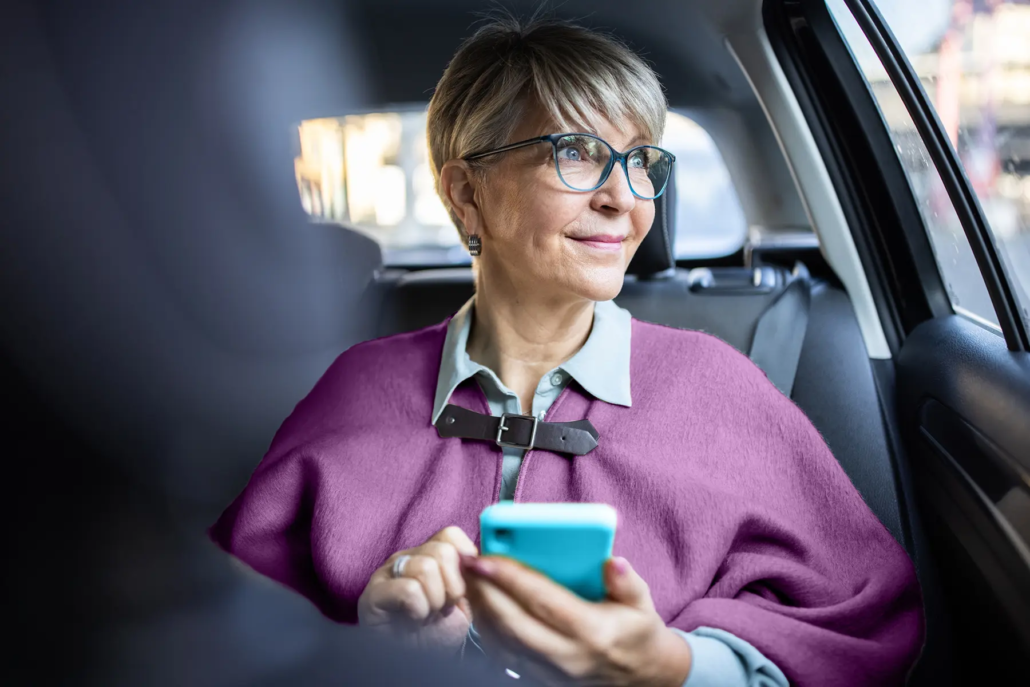 A senior woman sitting in the back seat of a car holding her smartphone and looking out the window with a happy expression.